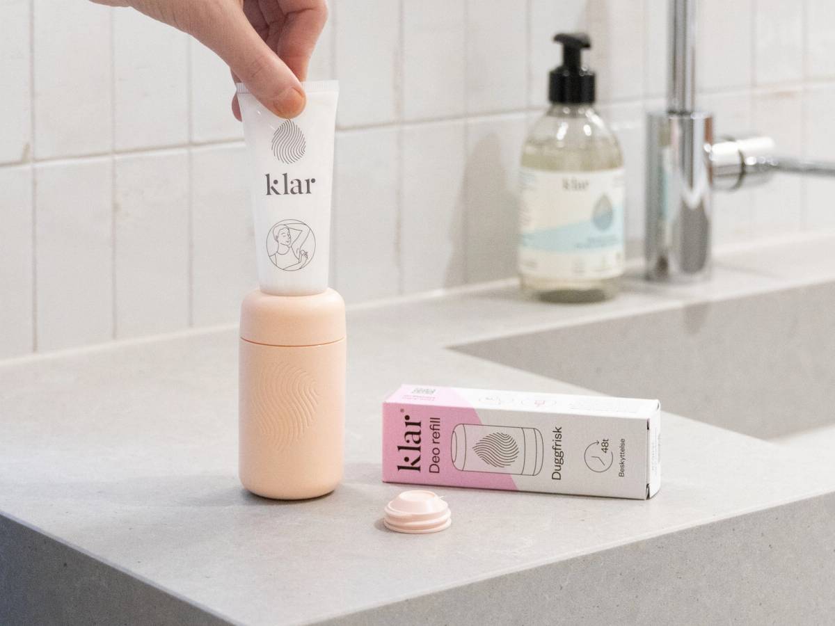 En person holder en hvit deodorant med etiketten 'klar' over en rosa og hvit deodorantboks på kjøkkenbenken.