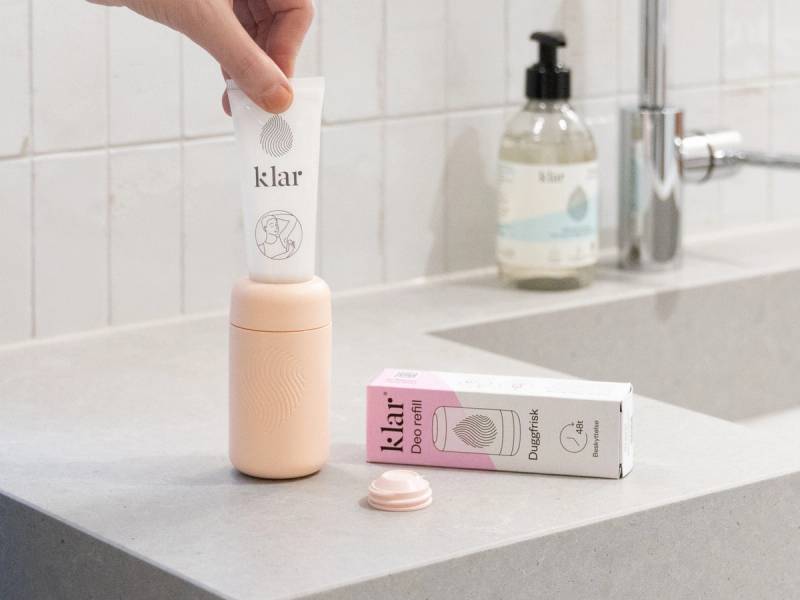 En person holder en hvit deodorant med etiketten 'klar' over en rosa og hvit deodorantboks på kjøkkenbenken.