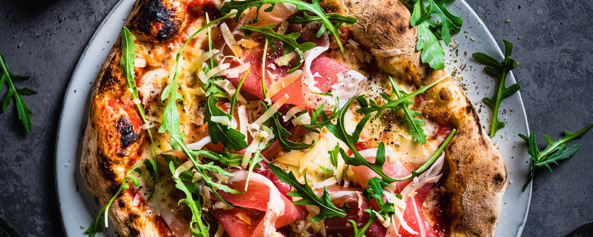 En pizza med skinke, ruccola og ost på en hvit tallerken.