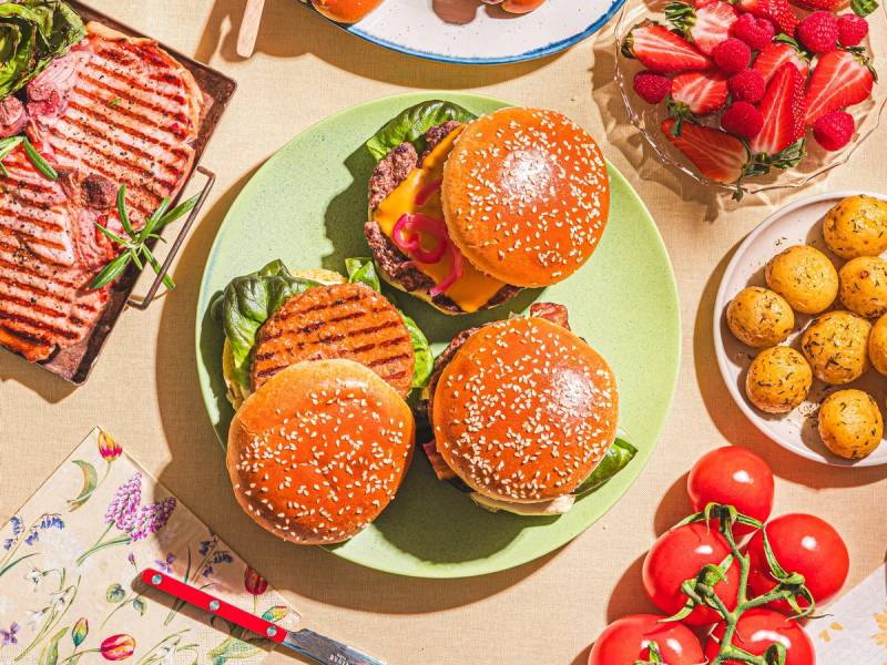 Tre burgere med sesongens grønnsaker på en grønn tallerken, omgitt av frukt og snacks på bordet.