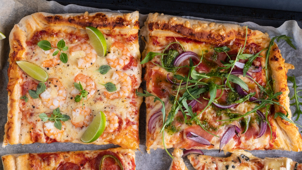 To store stykker langpannepizza med ost, skinke, grønnsaker og urter, delt i fire deler, på et stekebrett.