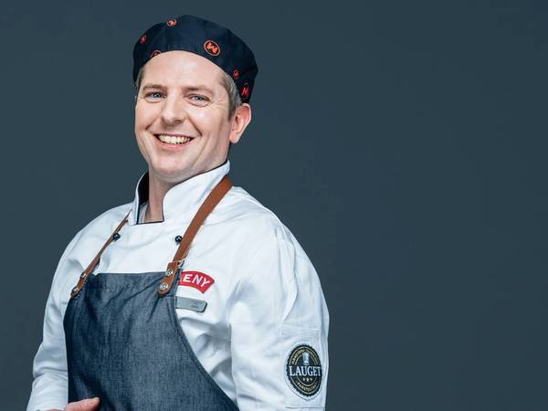 En person iført kokkeuniform og forkle, med armene i kors, mot en mørk bakgrunn.
