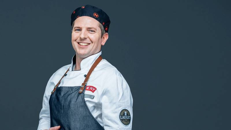 En person iført kokkeuniform og forkle, med armene i kors, mot en mørk bakgrunn.