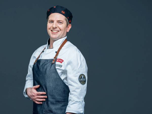 En person iført kokkeuniform og forkle, med armene i kors, mot en mørk bakgrunn.