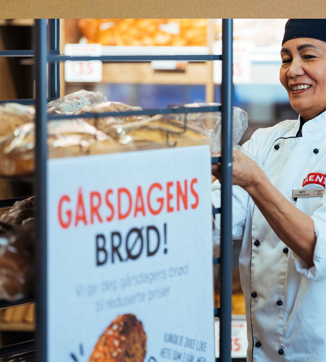 En baker i hvit kokkedrakt står ved en bakerihylle med brød, med et skilt som oppfordrer til å redusere matsvinn.