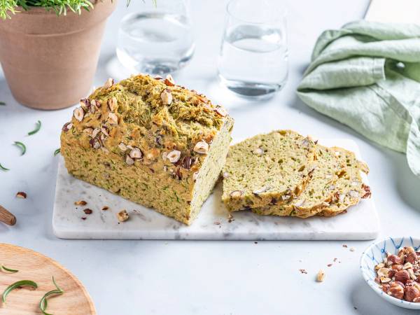 Et stykke brød med squash og nøtter på en hvit skjærefjøl, omgitt av glass og planter