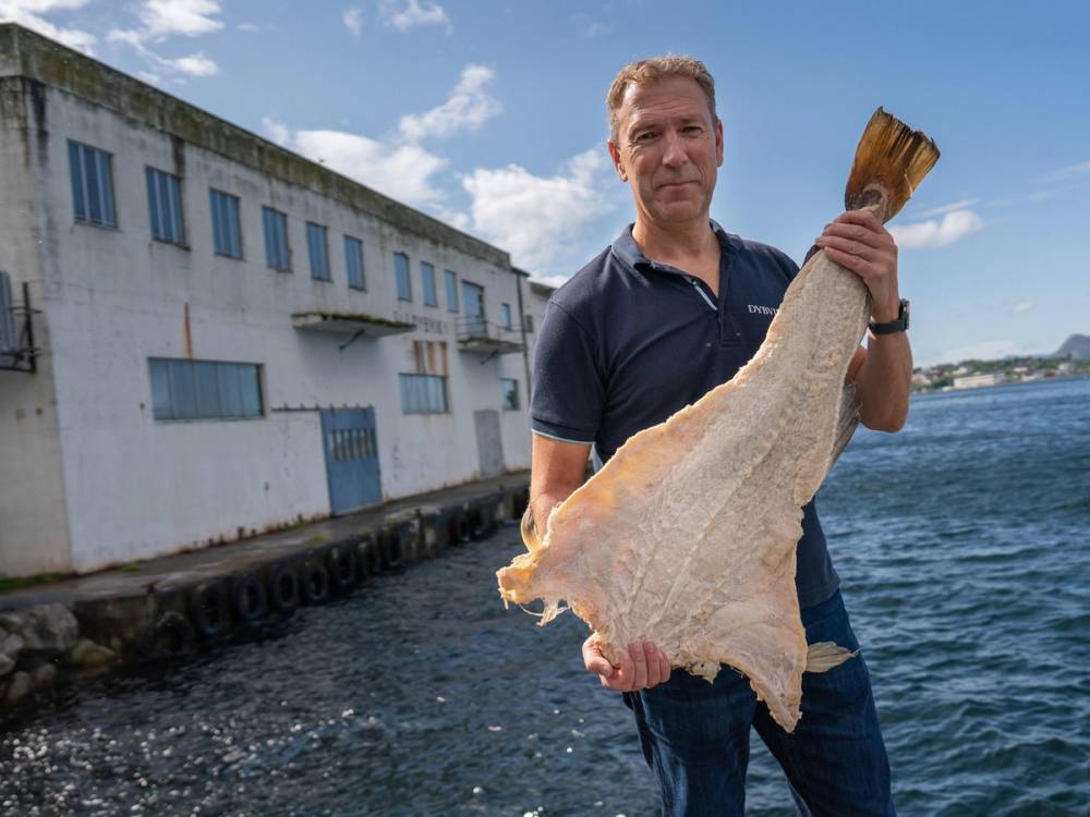 Person som holder en stor fisk ved vannet med en bygning i bakgrunnen, trolig i Møre-regionen.