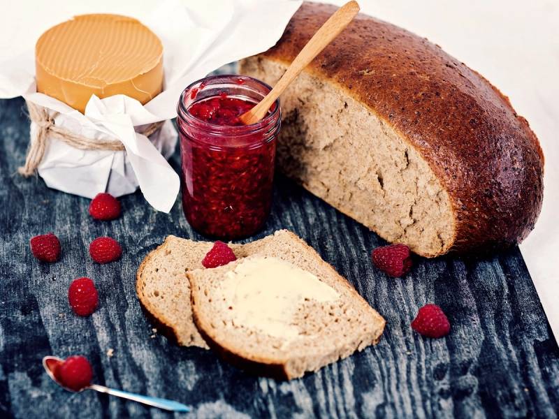Et brød med to skiver skåret av, en glass med rød syltetøy og en liten ost på et mørkt treplate, omgitt av noen bringebær.
