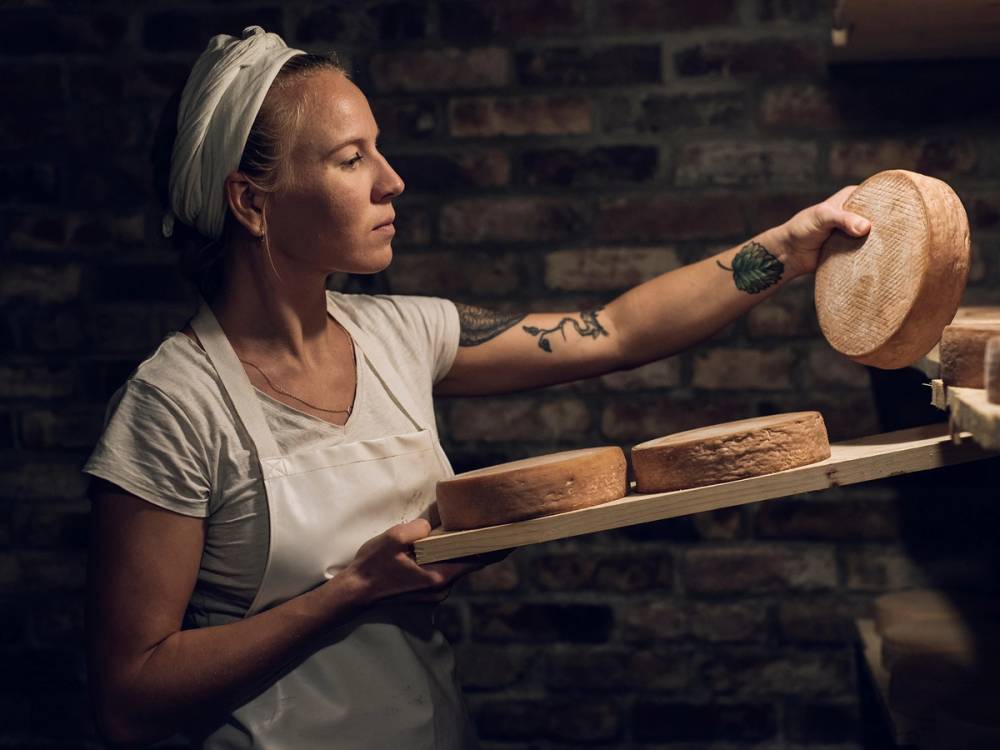 En person med tatoveringer på armen holder opp en stor ostebit i en ostebutikk eller et ysteri i Stavanger, Rogaland.
