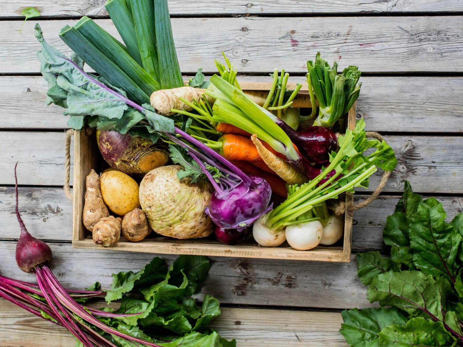 Et utvalg av rotgrønnsaker som gulrøtter, rødbeter, purre, og kålrot i en trekasse på en treplate.