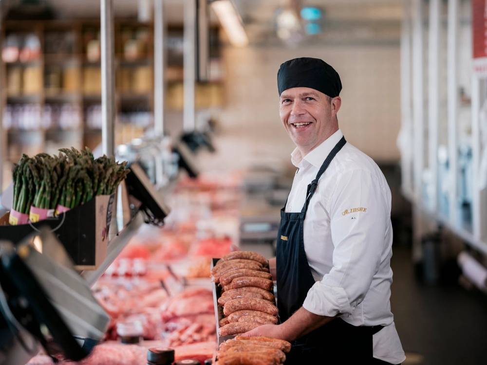 En person i hvit skjorte og svart forkle står bak en kjøttdisk i en butikk, med kjøttprodukter utstilt foran seg.