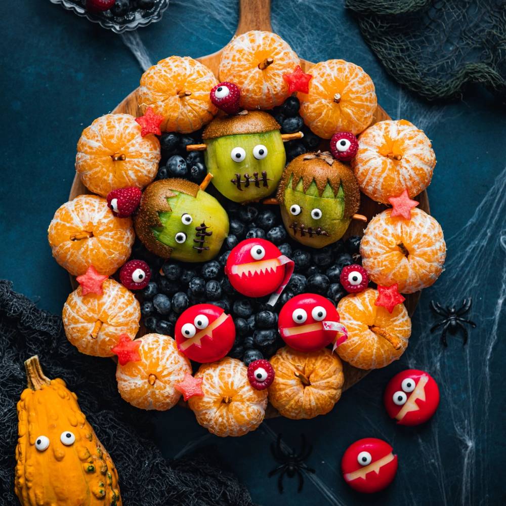 En Halloween-inspirert kakedekorasjon med små gresskar, monster-figurer og røde øyne, omgitt av små gresskar og svarte edderkopper på en mørk bakgrunn.