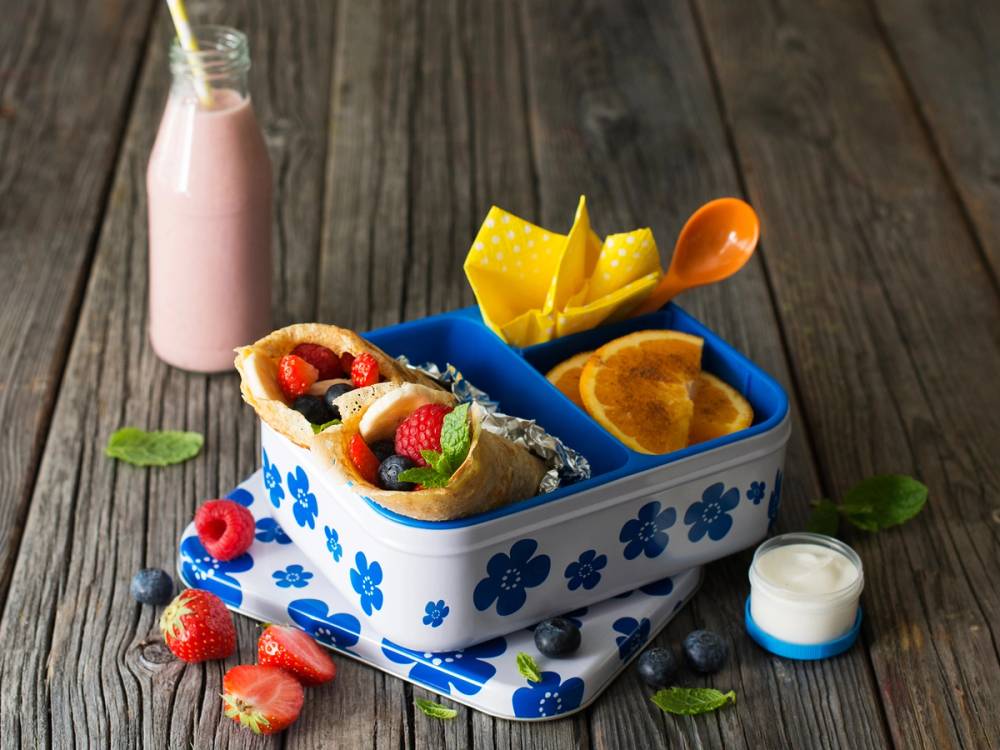 Grove pannekaker med bær og frukt i en matboks, sammen med en liten flaske med melk og en kopp med pommes frites på en treplate.