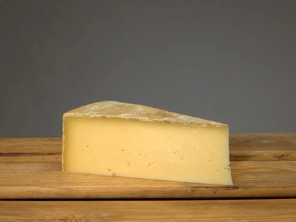 Et stykke ost på en trebordflate med en grå bakgrunn, trolig en del av et ostefat eller en matopplevelse.