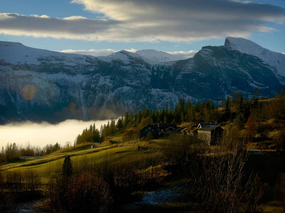 Utsikt over fjell og innsjø med små hytter i forgrunnen, omgitt av skog og fjell i bakgrunnen.