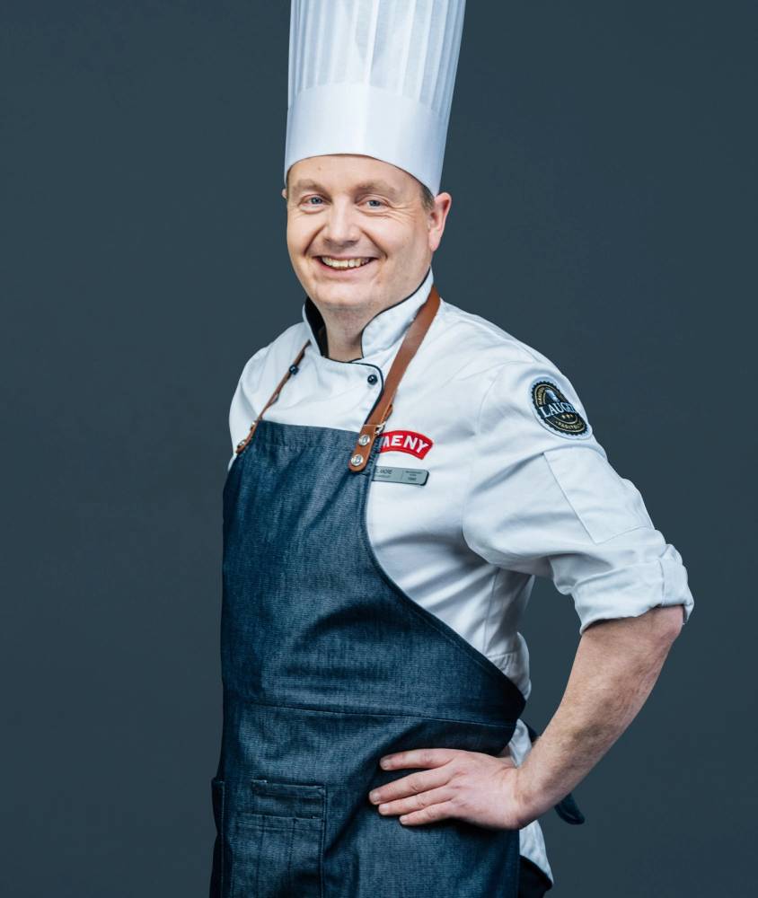 Kokk i hvit kokkejakke og blå forkle med kokkehatt, står med armene i siden mot en mørk bakgrunn.
