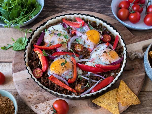 En tacopai med kjøttdeig, egg, cherrytomater, paprika og salat på et rundt fat, omgitt av tomater og tortillachips.