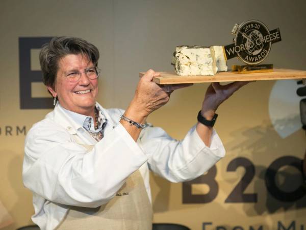 En person holder opp en treplate med en stor ost og en pokal på en premieutdeling, med en bakgrunn som viser tekst og logoer.