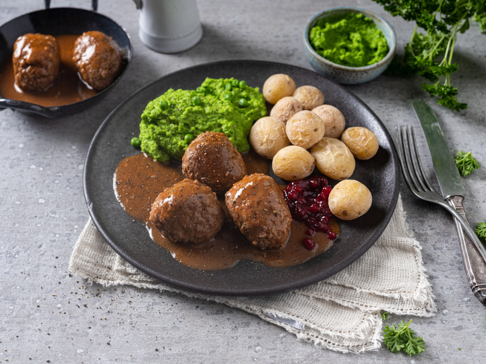 En tallerken med kjøttboller, poteter, brokkoli og tyttebær, plassert på et bord med bestikk og grønt dekorasjon rundt.