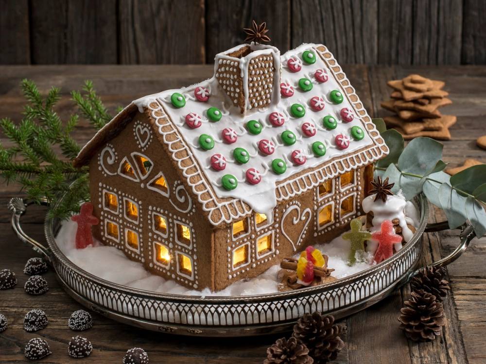Et pyntet pepperkakehus med lysende vinduer og dekorasjoner på et fat, omgitt av julepynt og granbar.