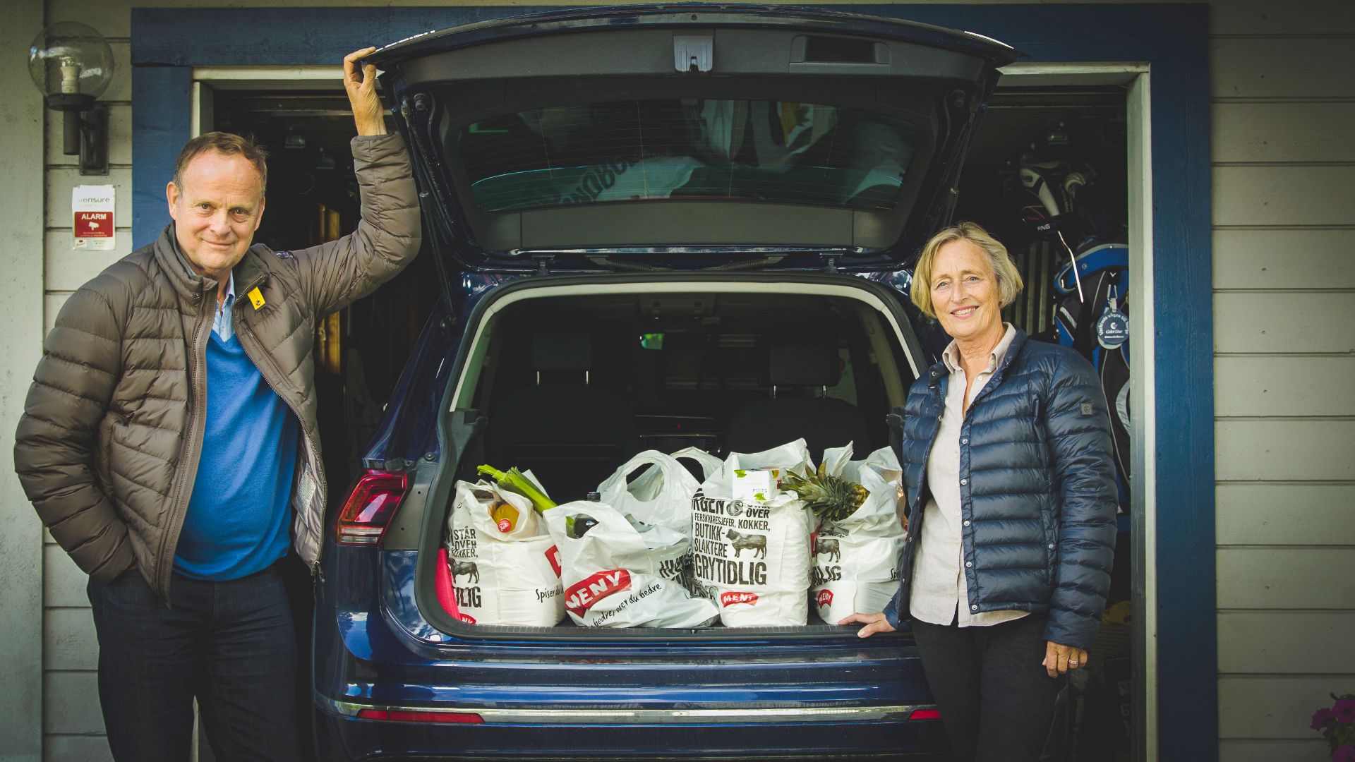 To personer står ved en åpen bil med varer i bagasjerommet, foran en blå husvegg.