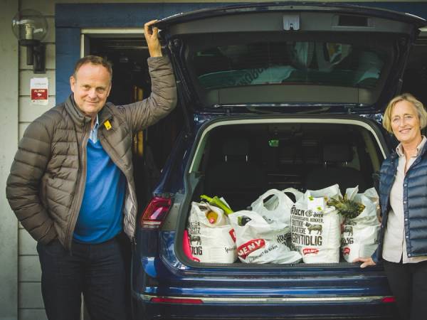 To personer står ved en åpen bil med varer i bagasjerommet, foran en blå husvegg.