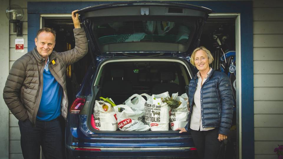 To personer står ved en åpen bil med varer i bagasjerommet, foran en blå husvegg.
