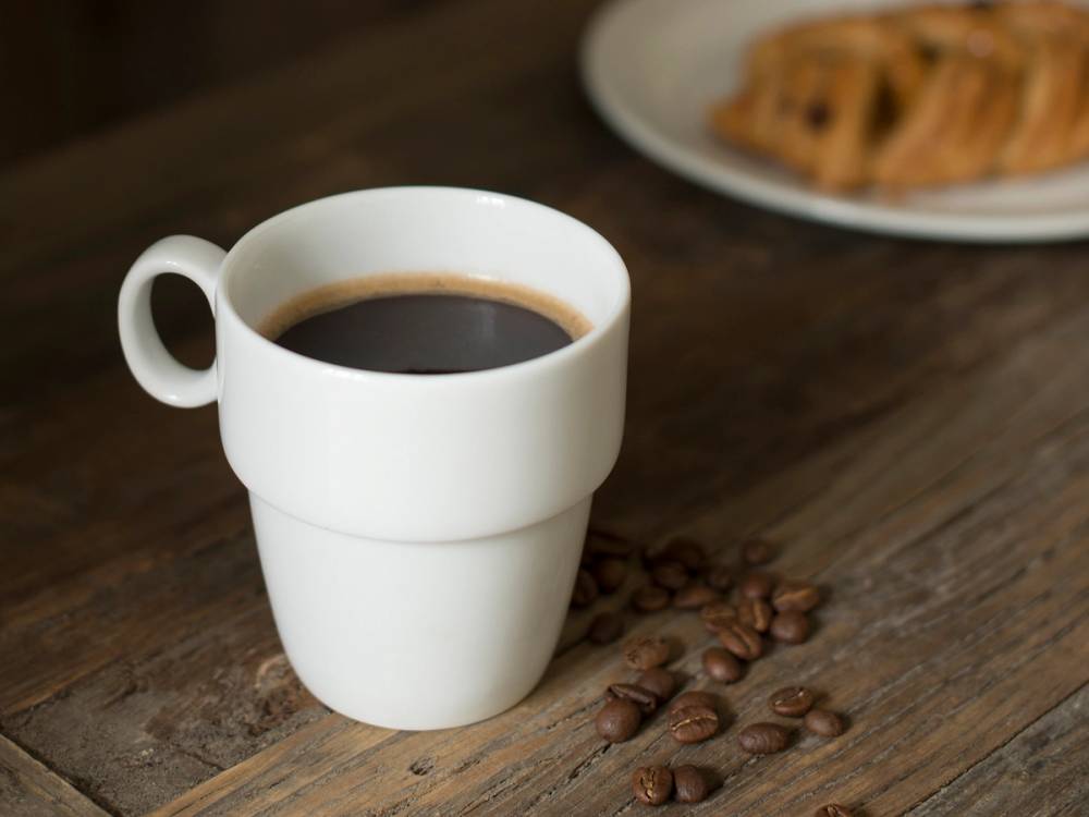 En hvit kaffekopp fylt med svart kaffe på et trebord med noen kaffebønner ved siden av.