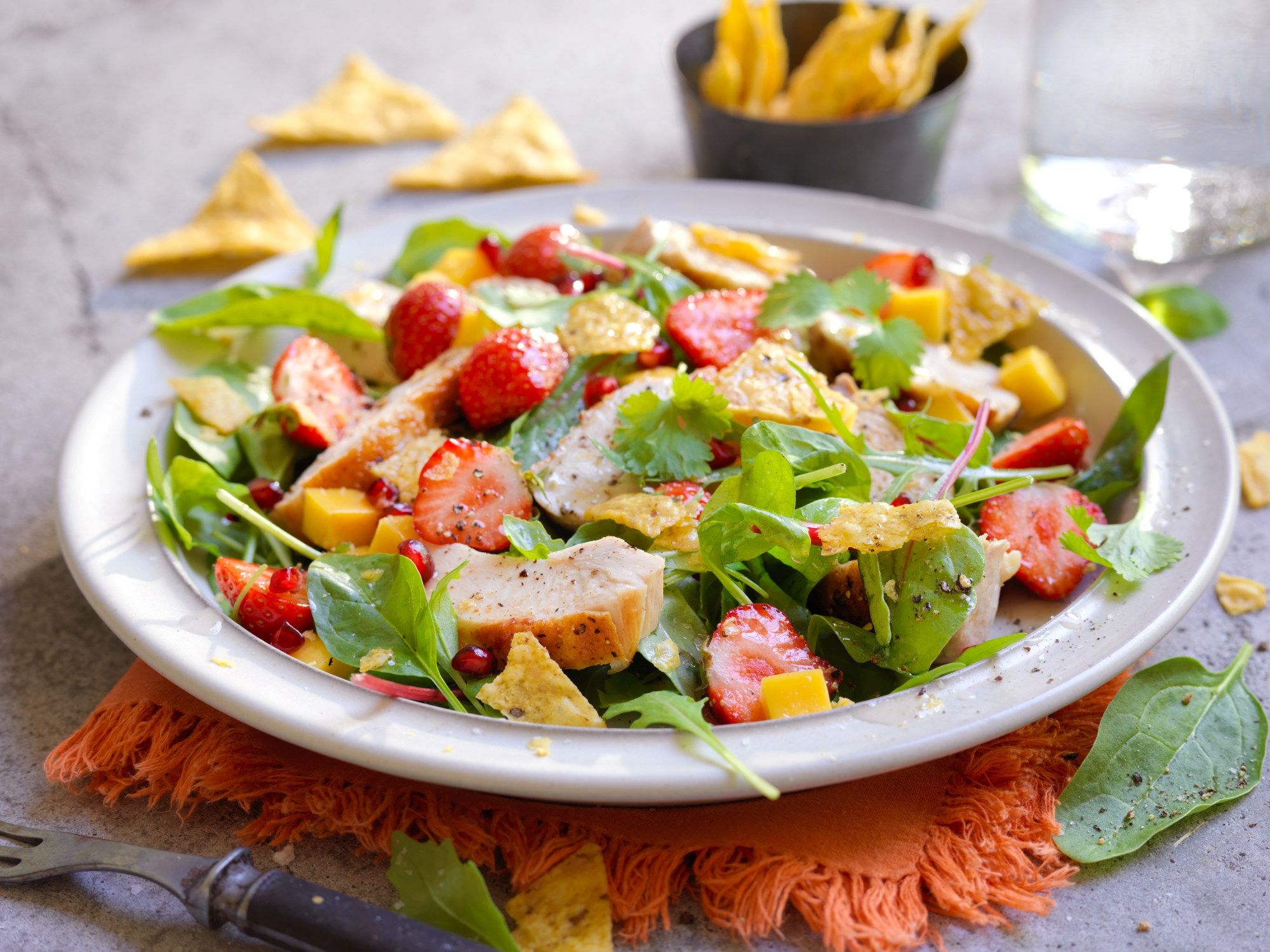 En frisk jordbærsalat med kyllingbiter og grønne blader på en hvit tallerken, med tortilla chips i bakgrunnen.