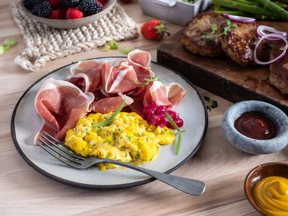 Spekemat med skinke, eggerøre og tilbehør på et fat, med en skive kjøtt og en liten skål med saus i bakgrunnen.