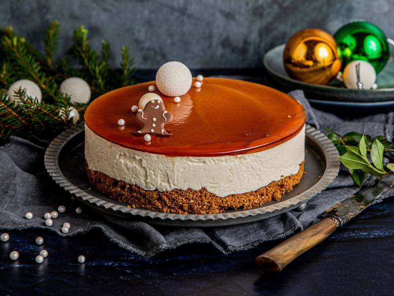 Ostekake med karamellisert topp og kjeksbunn, pyntet med små hvite og sølvfargede dekorasjoner, omgitt av julepynt og grønne grener.