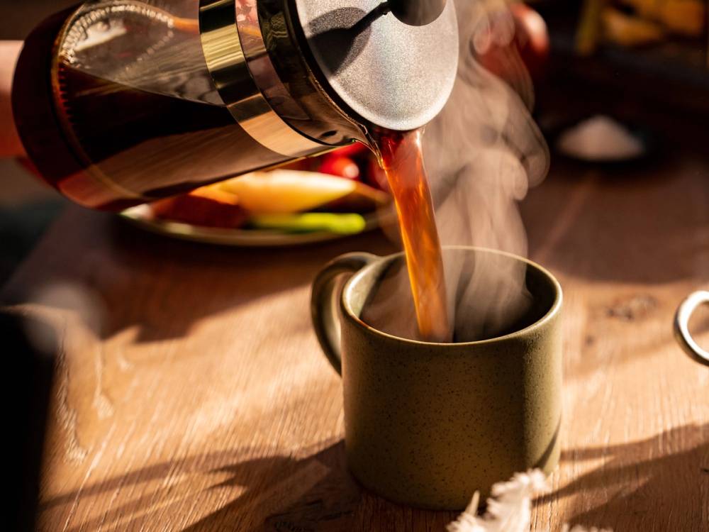 Kaffe helles fra en presskanne opp i en svart kopp på et trebord.