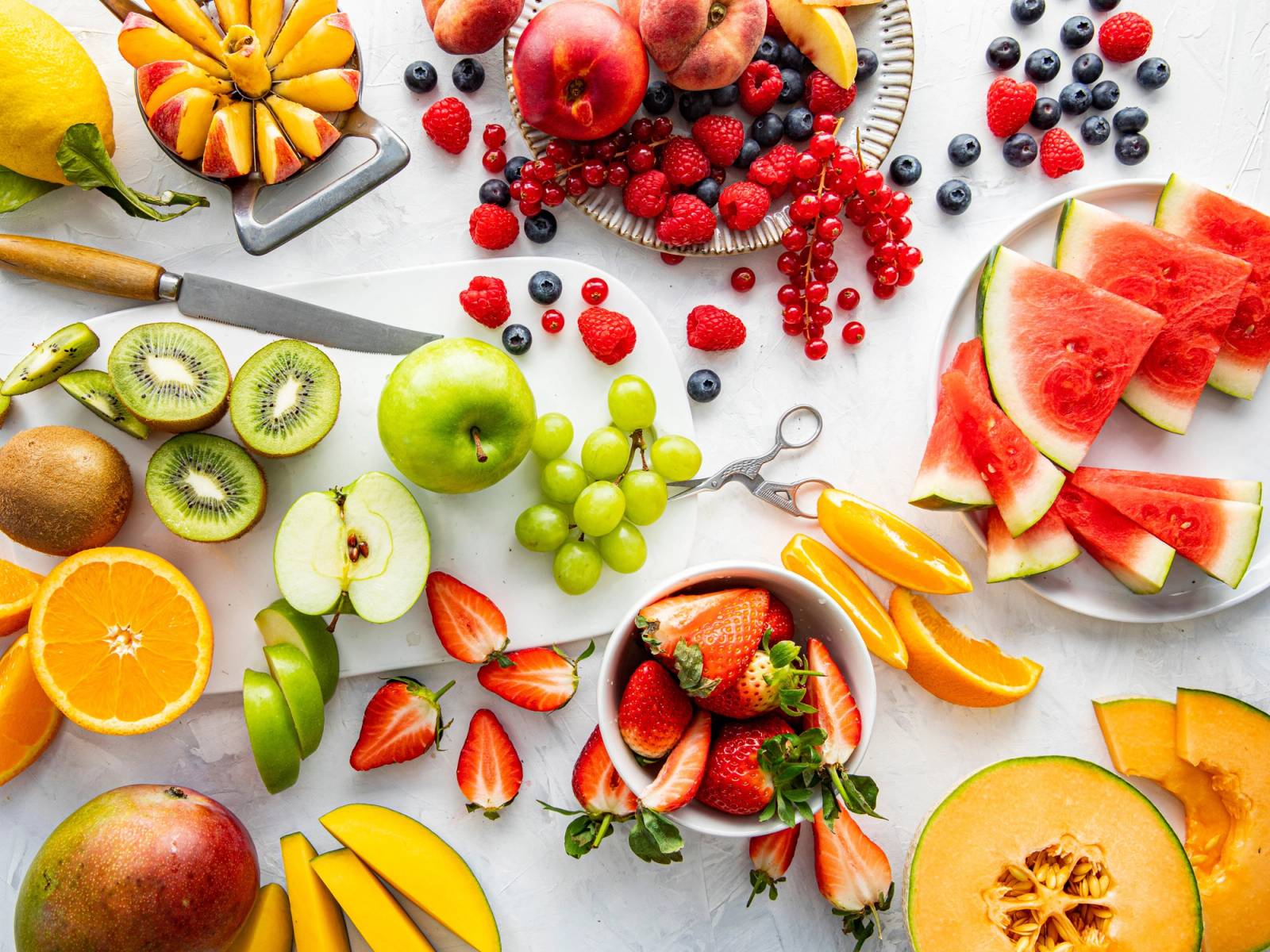 Fargerik oppskjæring av ulike frukter som vannmelon, kiwi, eple, druer, jordbær, appelsin og bær på et hvitt bord.