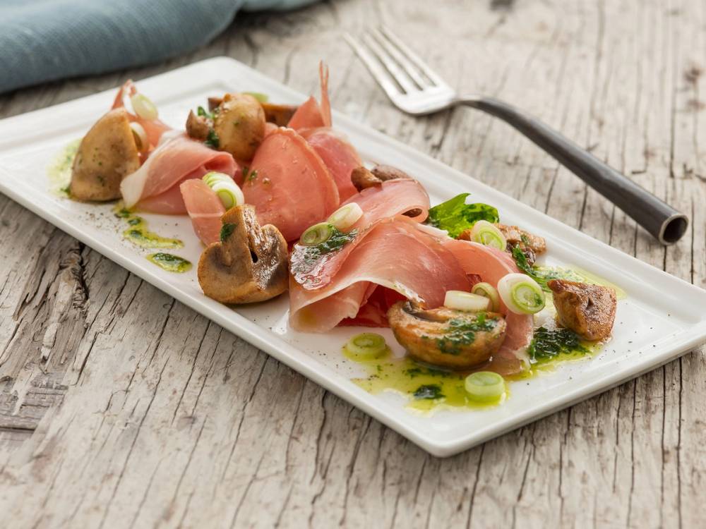 Spekeskinke med sopp og persilleolje servert på en hvit tallerken med grønne løk og sopp, på et treverkebord.