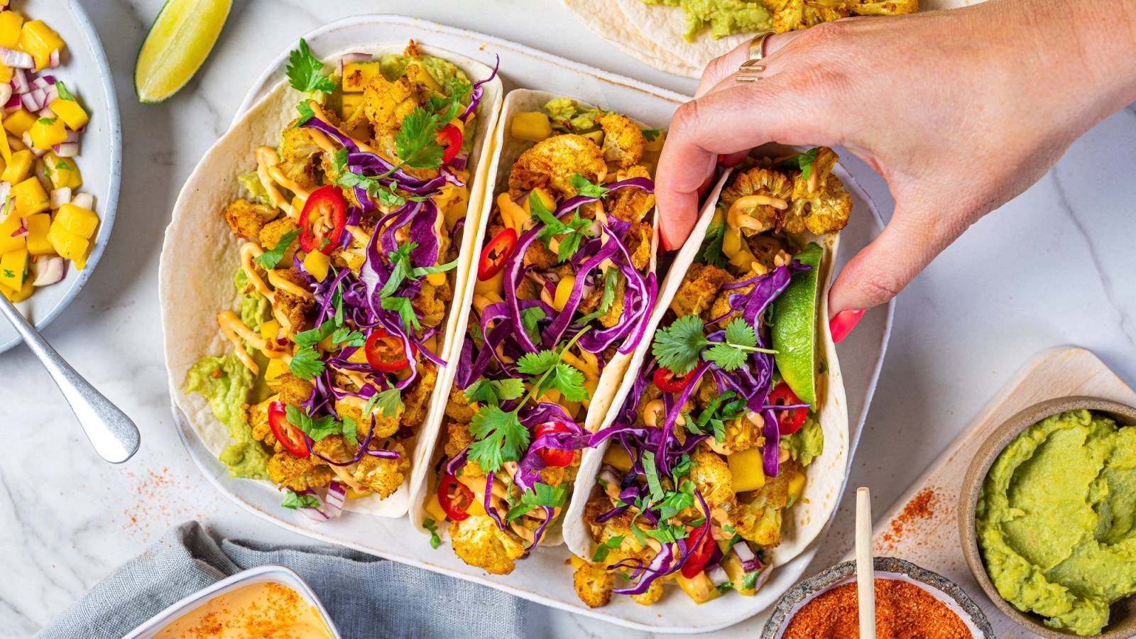 Tacos fylt med blomkål, kål og grønnsaker, med en hånd som tar en taco, omgitt av ulike tilbehør og guacamole.