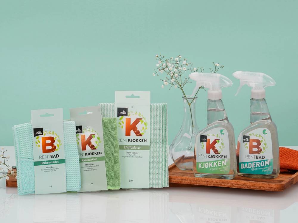 En rekke rengjøringsprodukter og sprayflasker står på et bord med en liten grein i en vase ved siden av.