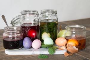 Glasskrukker med fargede væsker og fargede egg rundt på en hvid overflate, som kan tyde på kunstnerisk arbeid med å blåse egg.