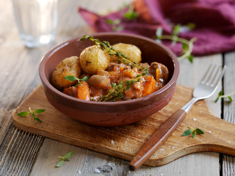 En bolle med boeuf bourguignon med kjøttboller, grønnsaker og urter, servert på et trebord med en glass vann og bestikk i nærheten.
