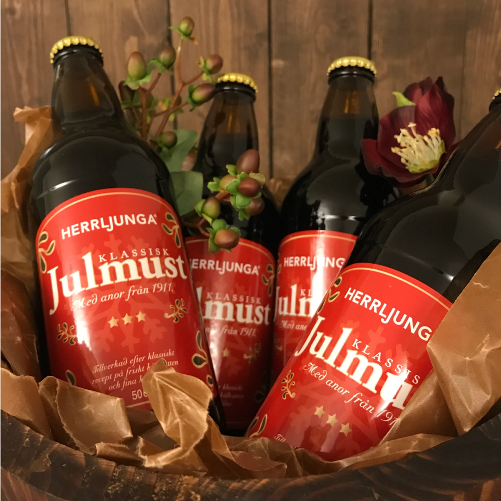 Flere flasker med julemust med røde etiketter og juledelementer rundt, plassert i en kurv.