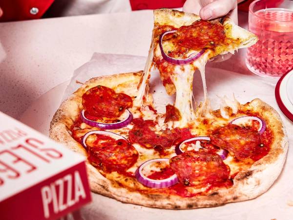 En person tar et stykke pepperoni-pizza med løk på en tallerken, med en pizzabit i hånden og en rosa drikk i bakgrunnen.