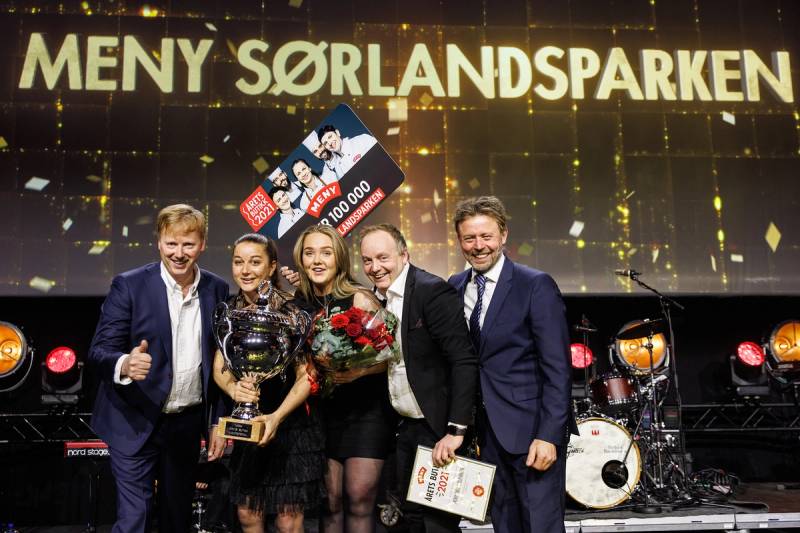 Fire personer står på scenen under en prisutdeling, med en stor skjerm som viser teksten 'MENY SØRLANDSPARKEN' i bakgrunnen.