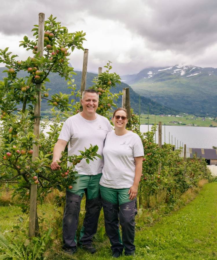 To personer står i en frukthage med trær og fjell i bakgrunnen, under en overskyet himmel.