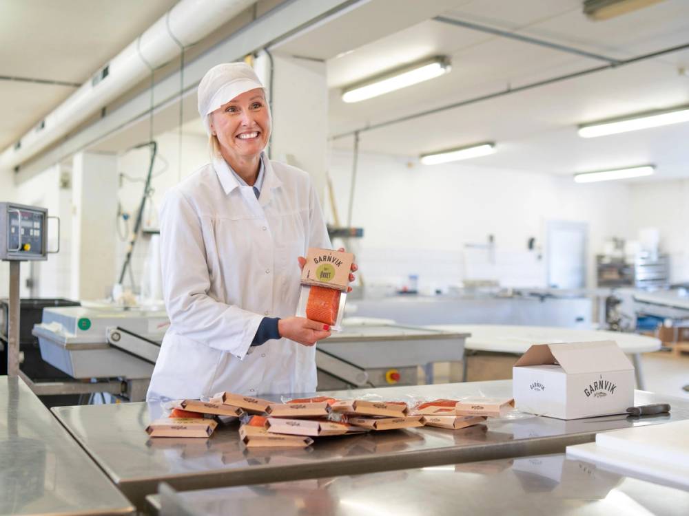 Person i hvit arbeidsdress pakker ferdiglagde matpakker i et industrielt kjøkken.