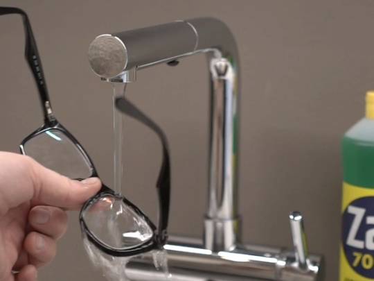 En hånd holder et par briller under en vannkran med Zalo-vaskemiddel i bakgrunnen.