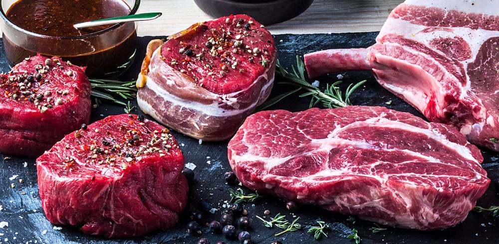 Ulike stykningsdeler av rått kjøtt, inkludert biffer og ribbe, på en mørk stekeplate med krydder og urter.