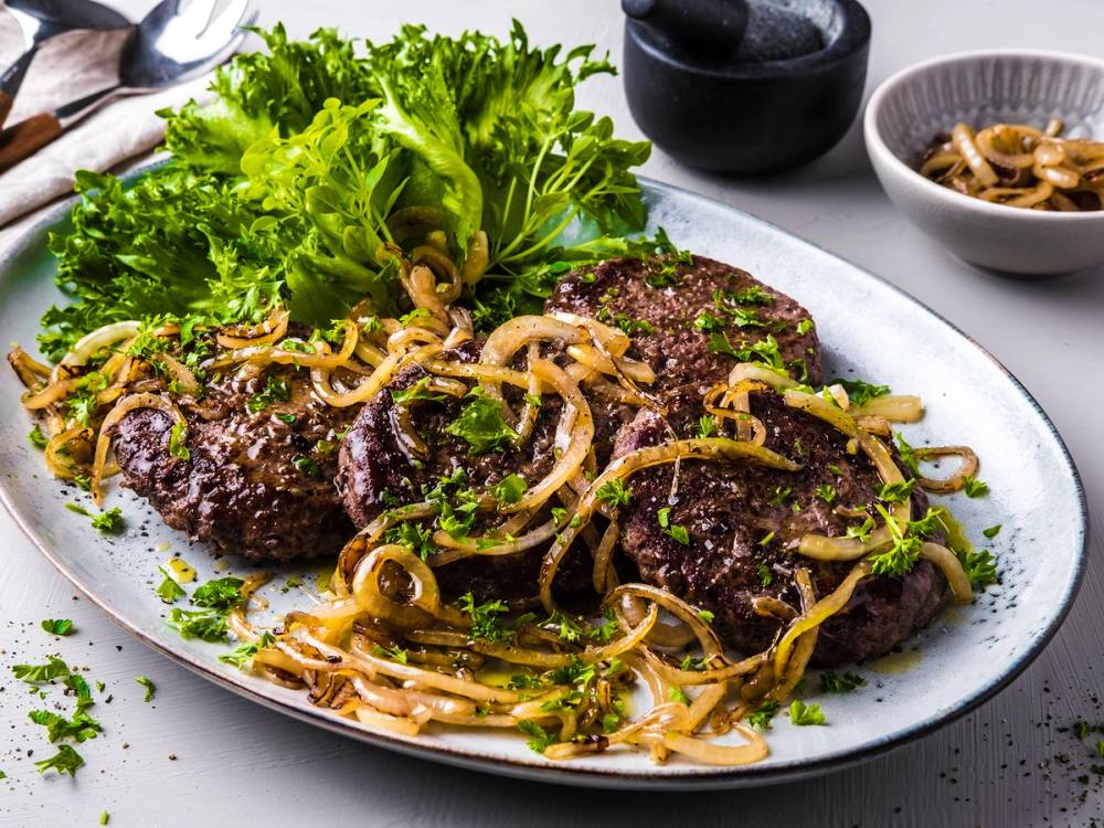 Karbonader med løk og grønn salat på en hvit tallerken, med en liten skål med sopp i bakgrunnen.