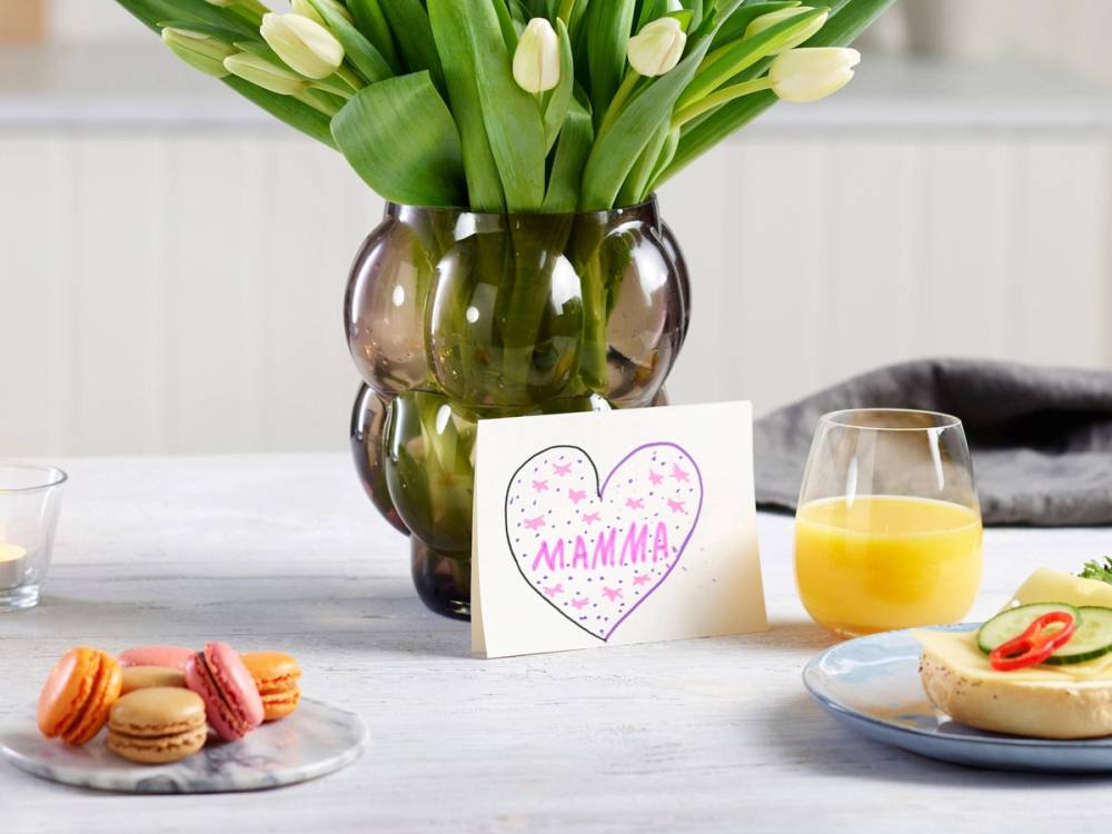 En vase med hvite tulipaner plassert på et bord sammen med en kort med teksten 'Mamma' og noen små kaker og en glass med appelsinjuice.