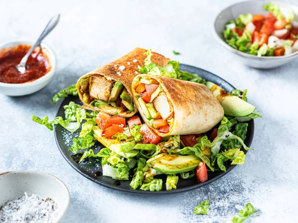 Tofuwrap med grønnsaker på en svart tallerken, omgitt av små skåler med saus og krydder, på et lyst bord.