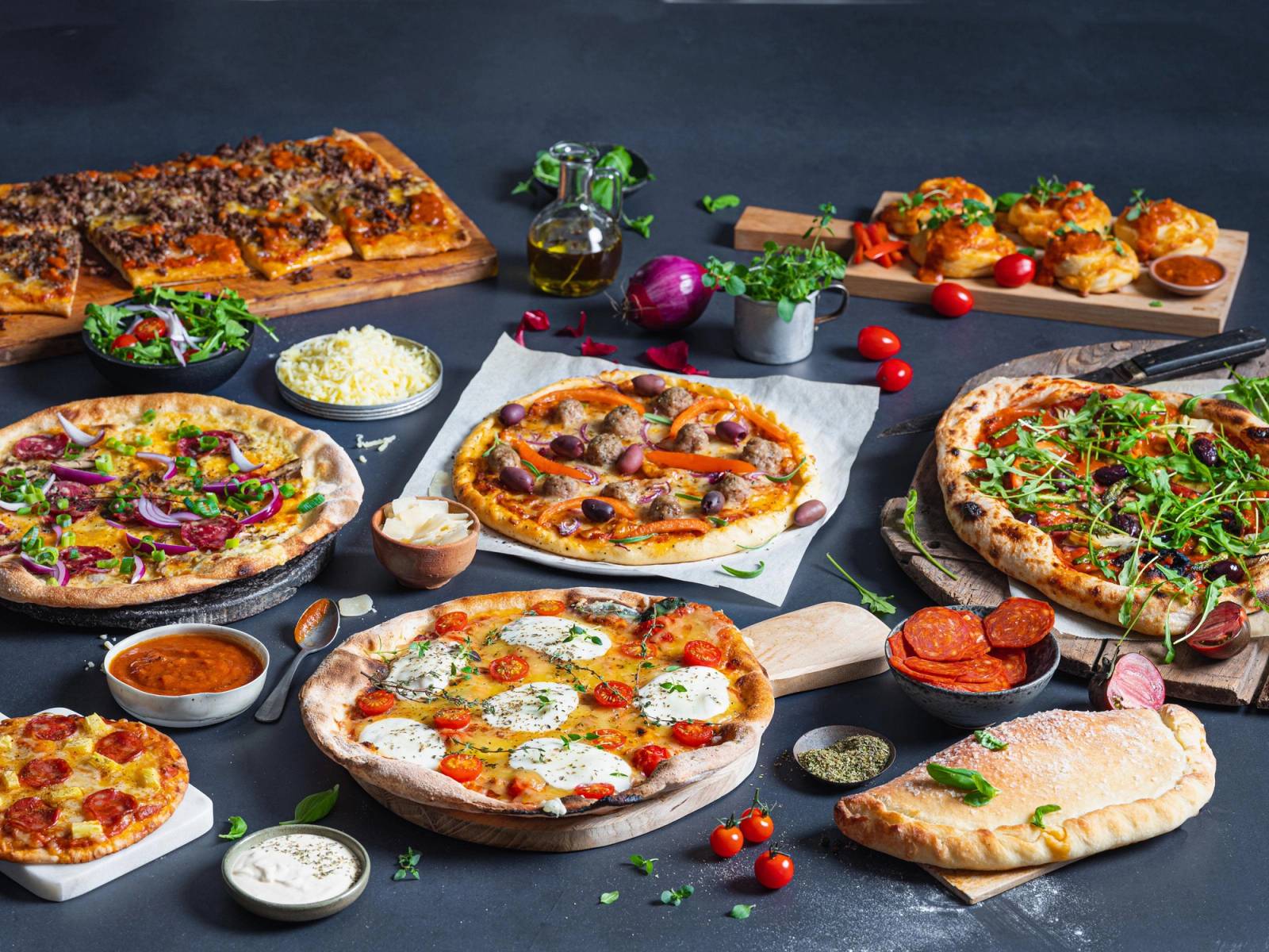 Et utvalg av pizzaer med ulike toppings på et bord, inkludert en pizza med skinke, en med grønnsaker og en med kjøtt, omgitt av små skåler med saus og tilbehør.