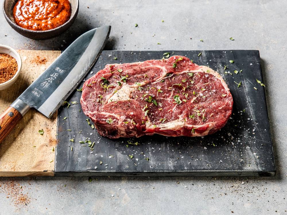 En rå entrecôte på et skjærebrett med krydder og en kniv ved siden av.